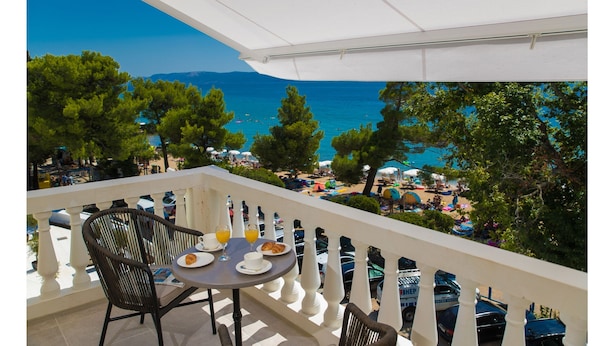 Un balcon avec une table préparée pour le petit-déjeuner, donnant sur une plage parsemée de parasols et animée par la présence de nombreuses personnes.