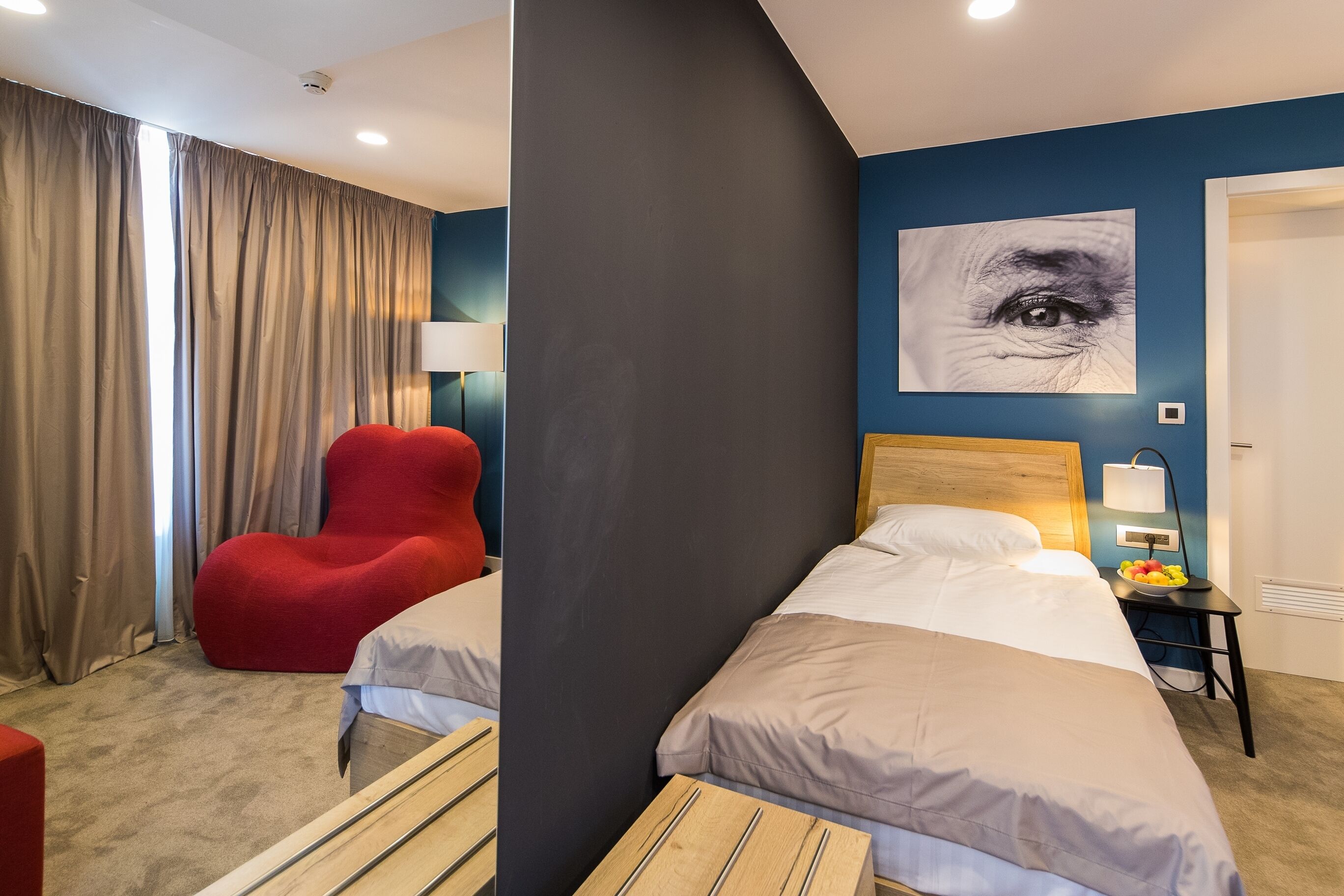 A hotel room with two beds, a red chair, a wall art of an eye, and a bedside table with a lamp and fruit bowl.