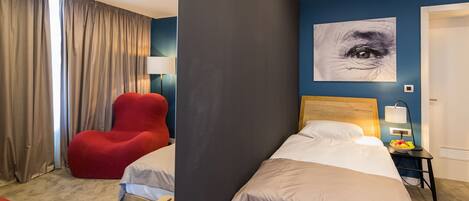 A hotel room with two beds, a red chair, a wall art of an eye, and a bedside table with a lamp and fruit bowl.