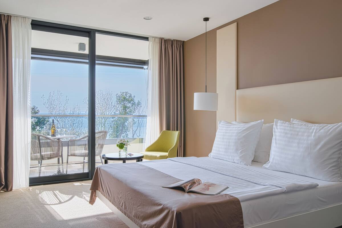 Une chambre d’hôtel avec un grand lit, une petite table sur laquelle est posé un vase, une chaise et un balcon avec vue.