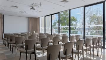 Ein Konferenzraum mit Reihen von Stühlen, einer Projektorleinwand und großen Fenstern, die einen Blick auf Bäume und ein Gebäude freigeben.
