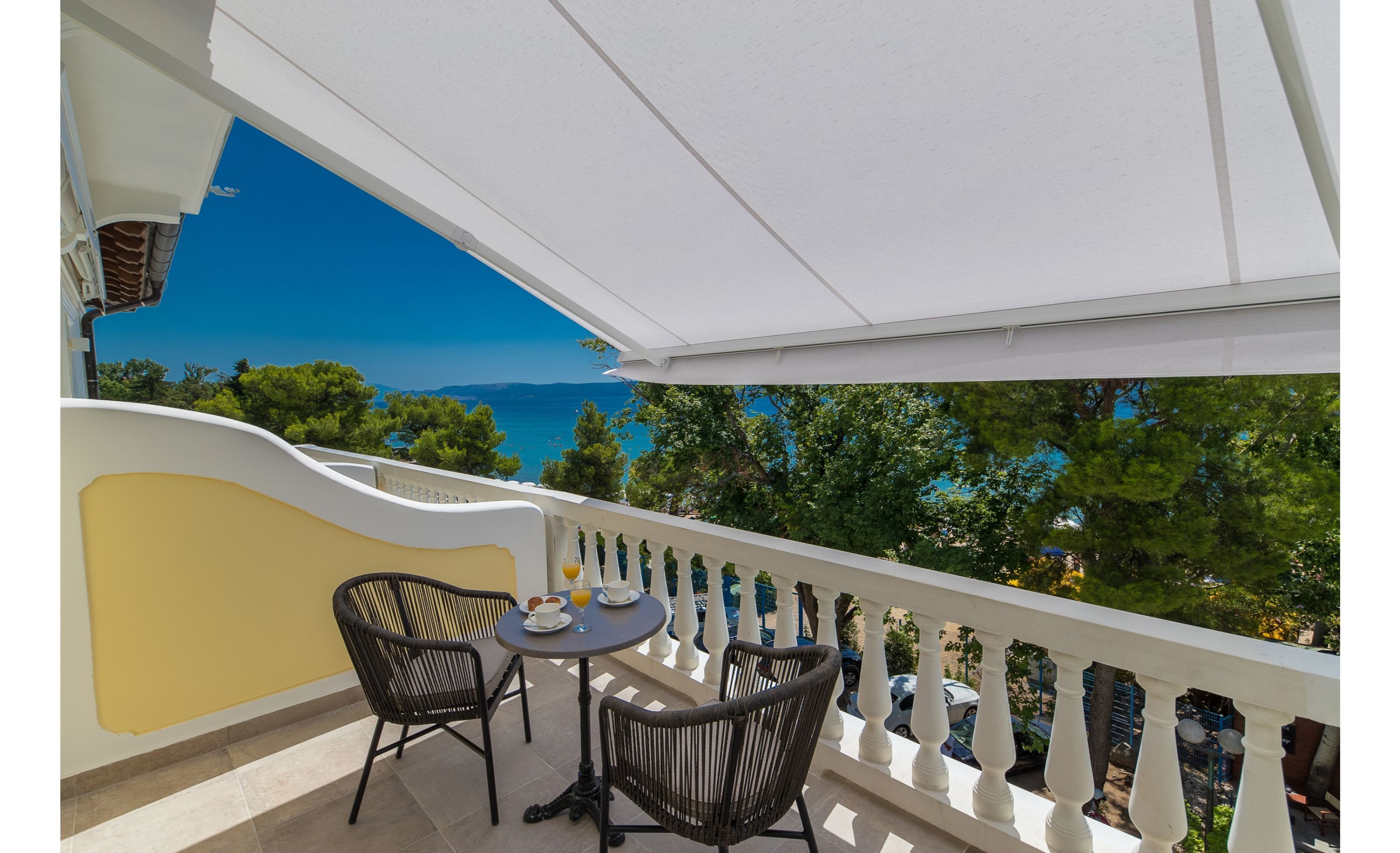 A balcony with a table and two chairs, overlooking a scenic view of the sea and trees.