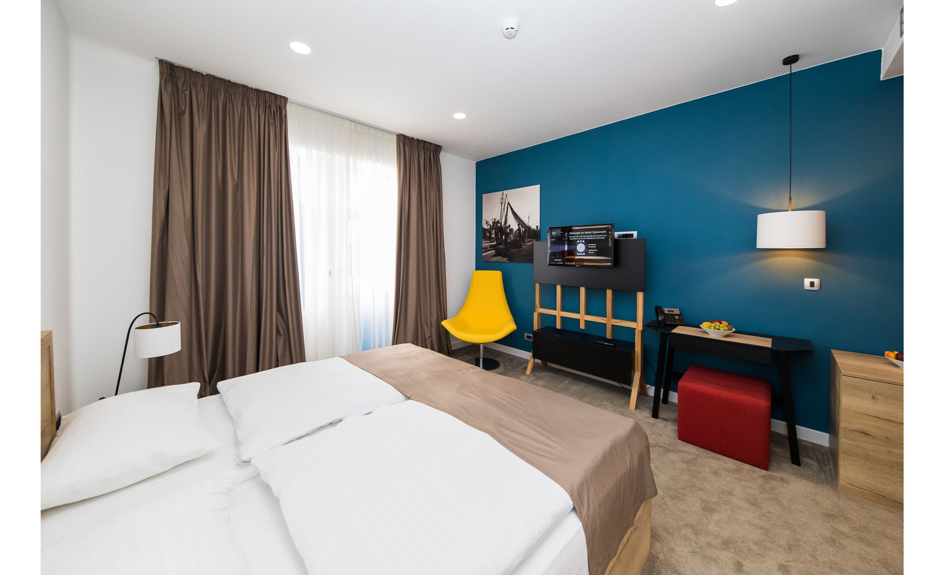 A hotel room with a bed, a desk, a chair, a TV, and a red ottoman.