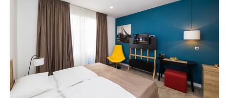 A hotel room with a bed, a desk, a chair, a TV, and a red ottoman.