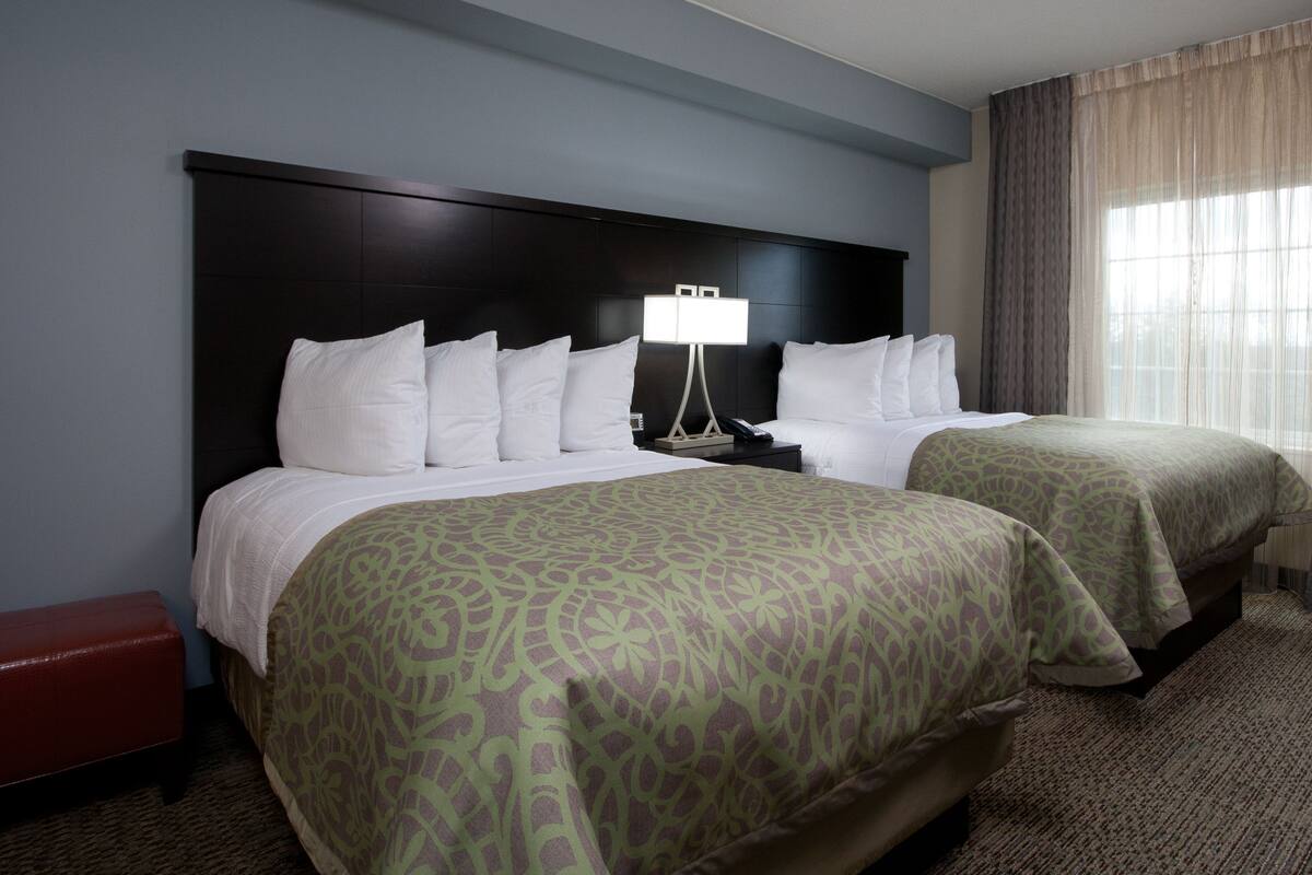 A hotel room with two beds, a nightstand with a lamp, a red bench, and a window with curtains.