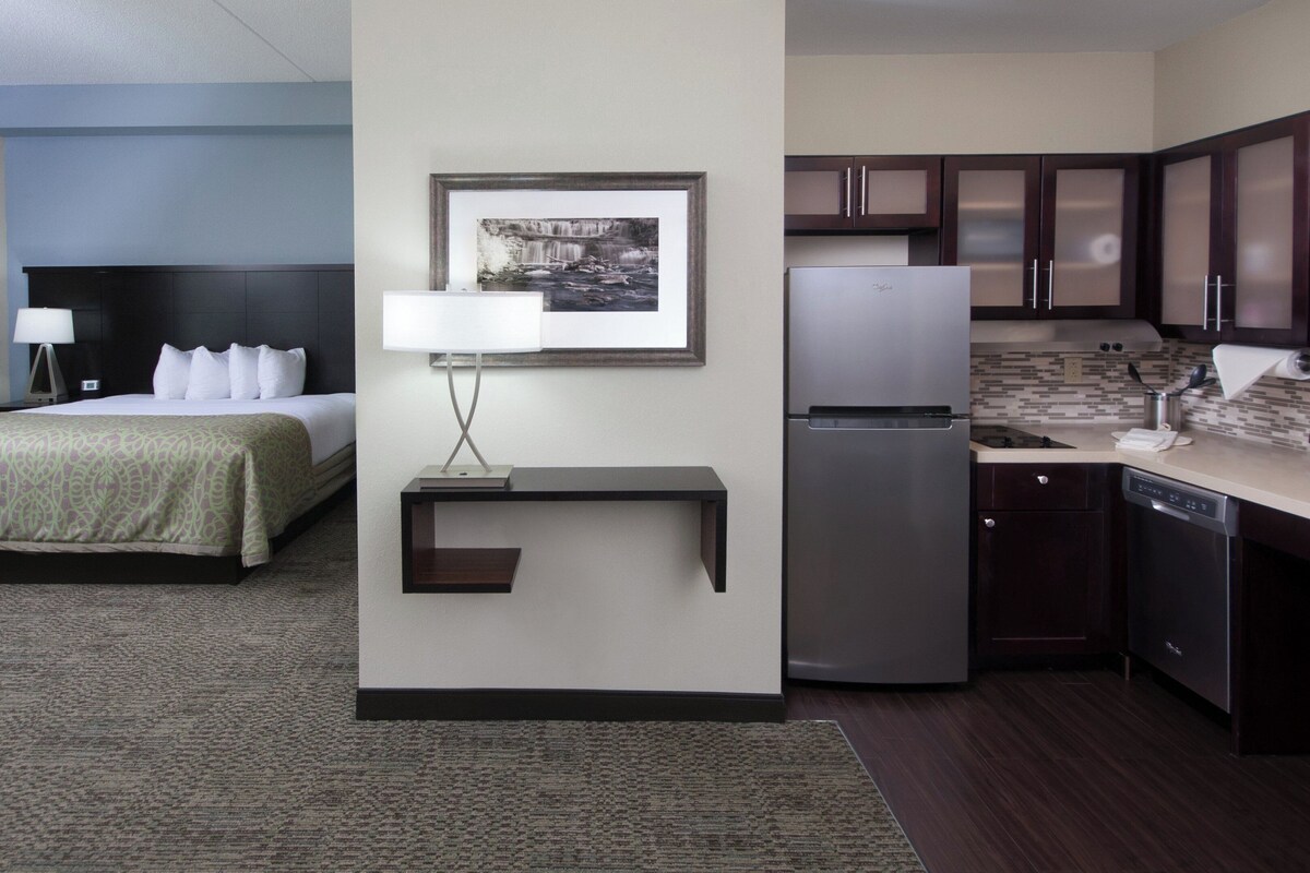 A hotel room with a bed, a desk with a lamp, a refrigerator, and a kitchen area.