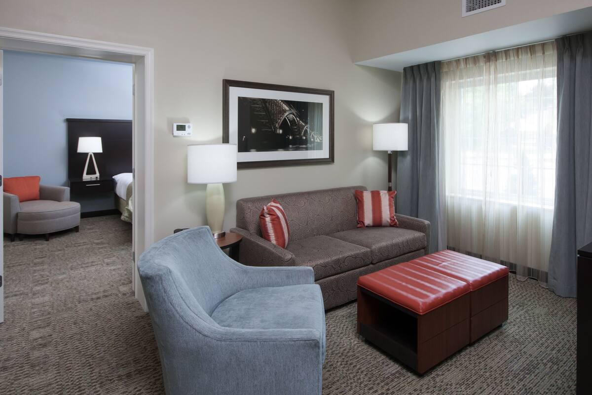 A hotel room with a grey armchair, a brown sofa with red cushions, a red ottoman, a framed picture on the wall, a lamp, and a window with grey curtains.