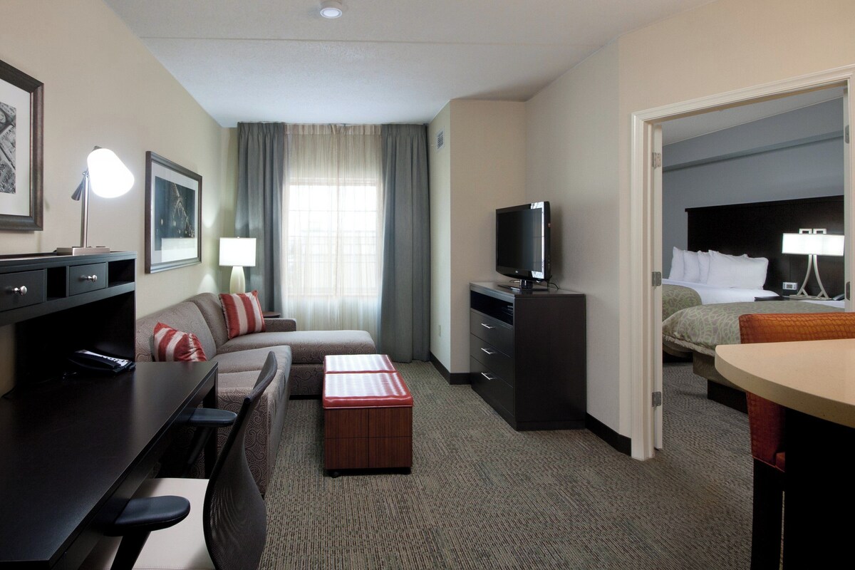 A hotel room with a sofa, a TV, a desk with a lamp, a small table, and a bed.
