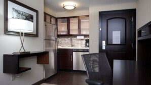 A compact kitchen with dark wooden cabinets, a stainless steel refrigerator, a dishwasher, and a black chair. - Staybridge Suites Buffalo-Amherst by IHG (Buffalo)