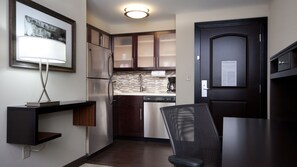 A compact kitchen with dark wooden cabinets, a stainless steel refrigerator, a dishwasher, and a black chair. - Staybridge Suites Buffalo-Amherst by IHG (Buffalo)