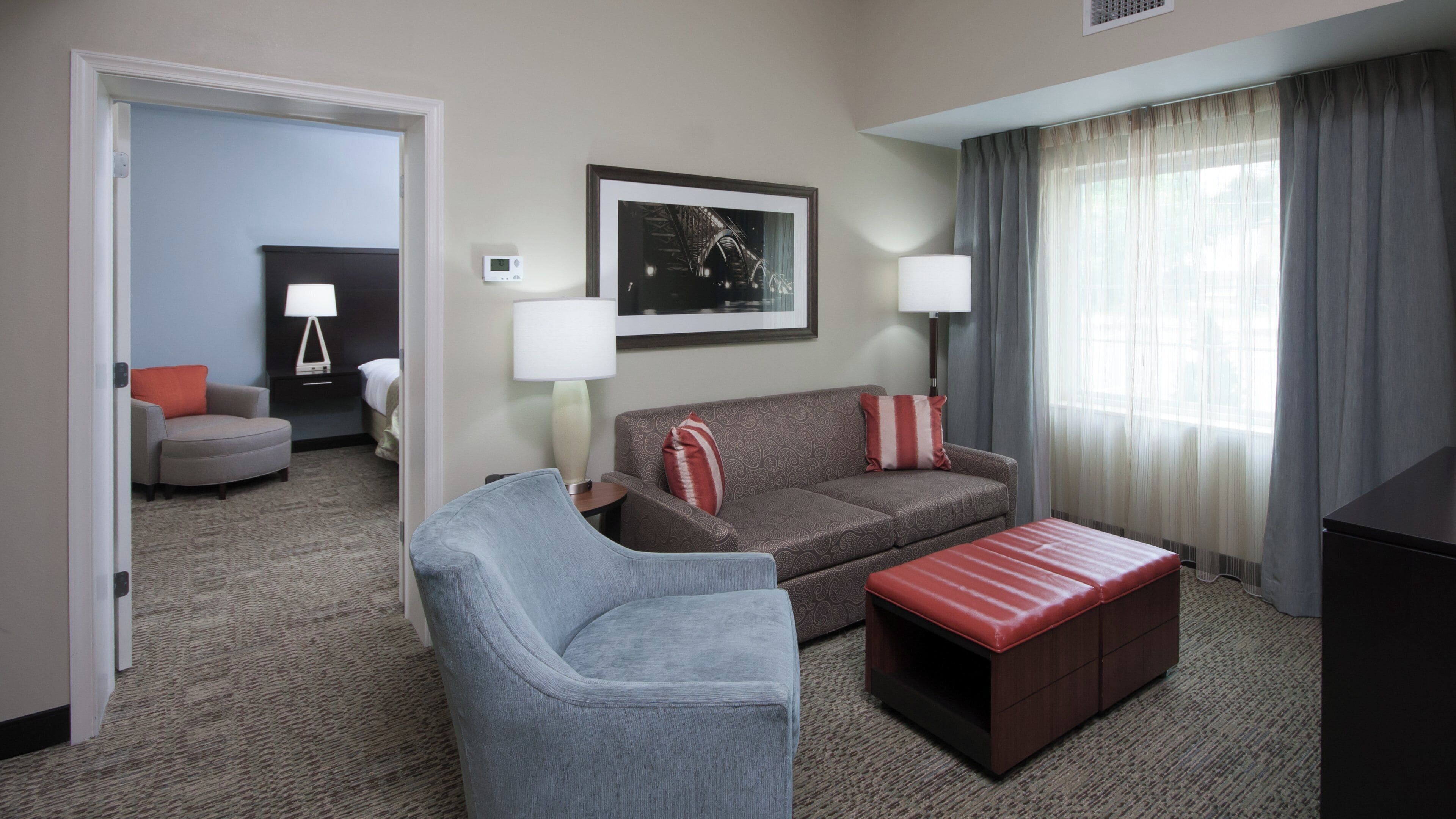A hotel room with a grey sofa, a red ottoman, a grey armchair, a framed picture on the wall, and a window with curtains.