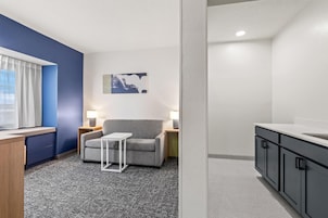 Una habitación de hotel moderna con un sofá gris, una mesita blanca, una pared de acento azul, un cuadro y una kitchenette.