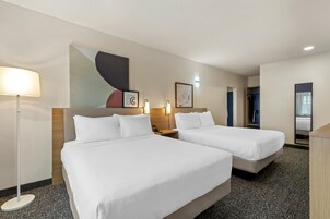 Habitación de hotel con dos camas, un escritorio, una lámpara y una obra de arte en la pared.