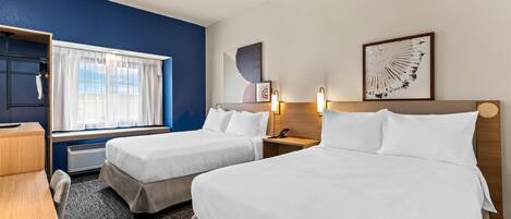 A hotel room with two beds, a desk, a chair, a window with curtains, a wall-mounted artwork, and a bedside table with a lamp.