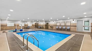 An indoor swimming pool with a tiled floor, a metal ladder, and a seating area with chairs and tables.