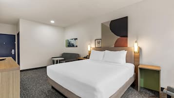 A hotel room with a bed, a desk, a chair, a small table, and a wall-mounted lamp.