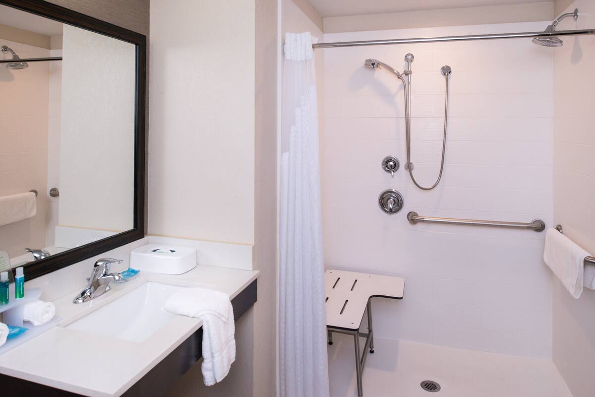 A bathroom with a sink, mirror, shower, and a small chair.