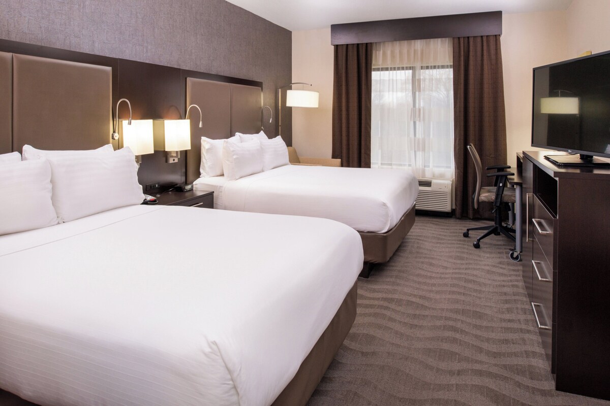 A hotel room with two beds, a desk, a television, and a large window with curtains.