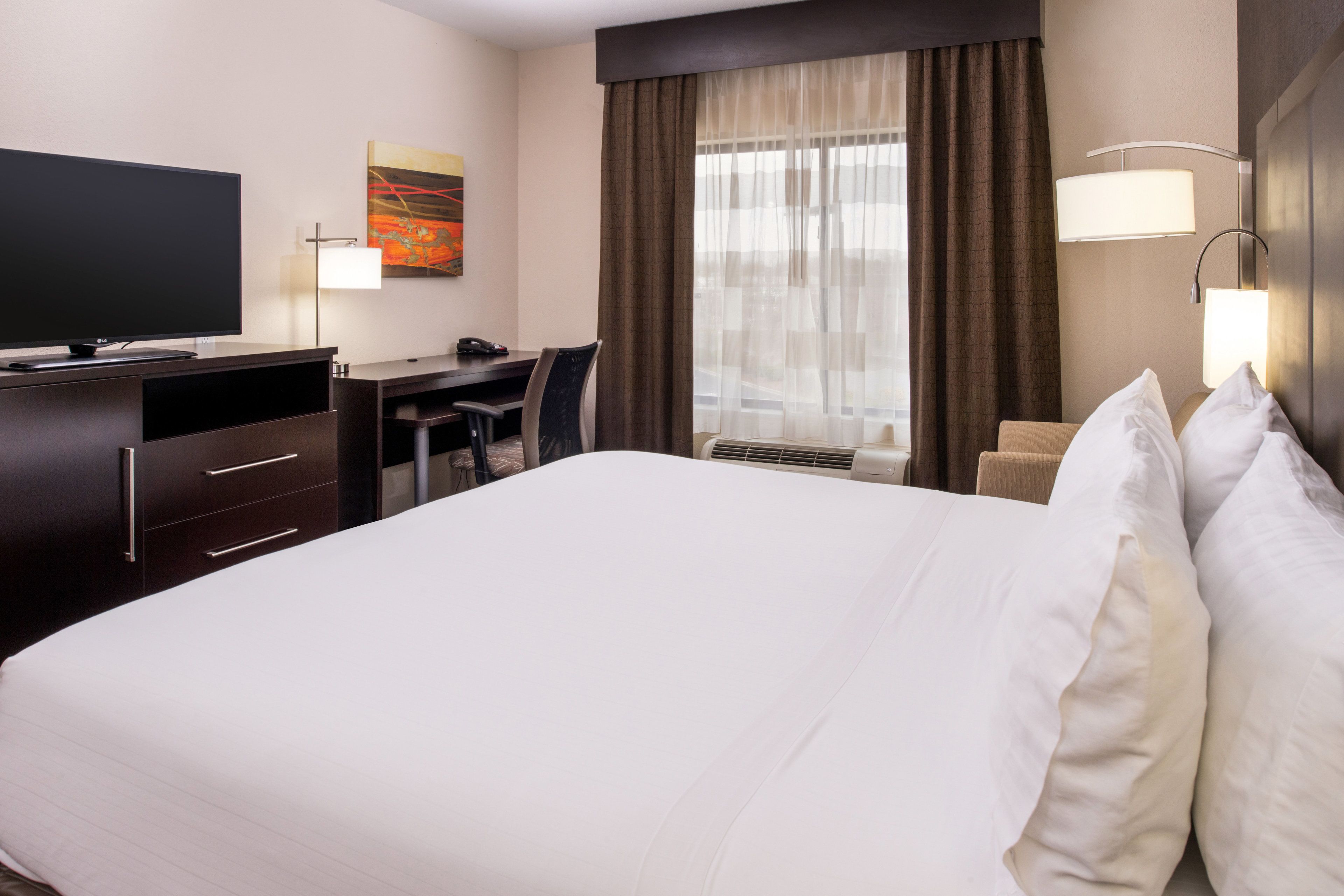 A hotel room with a large bed, a desk, a chair, a television, and a window with curtains.