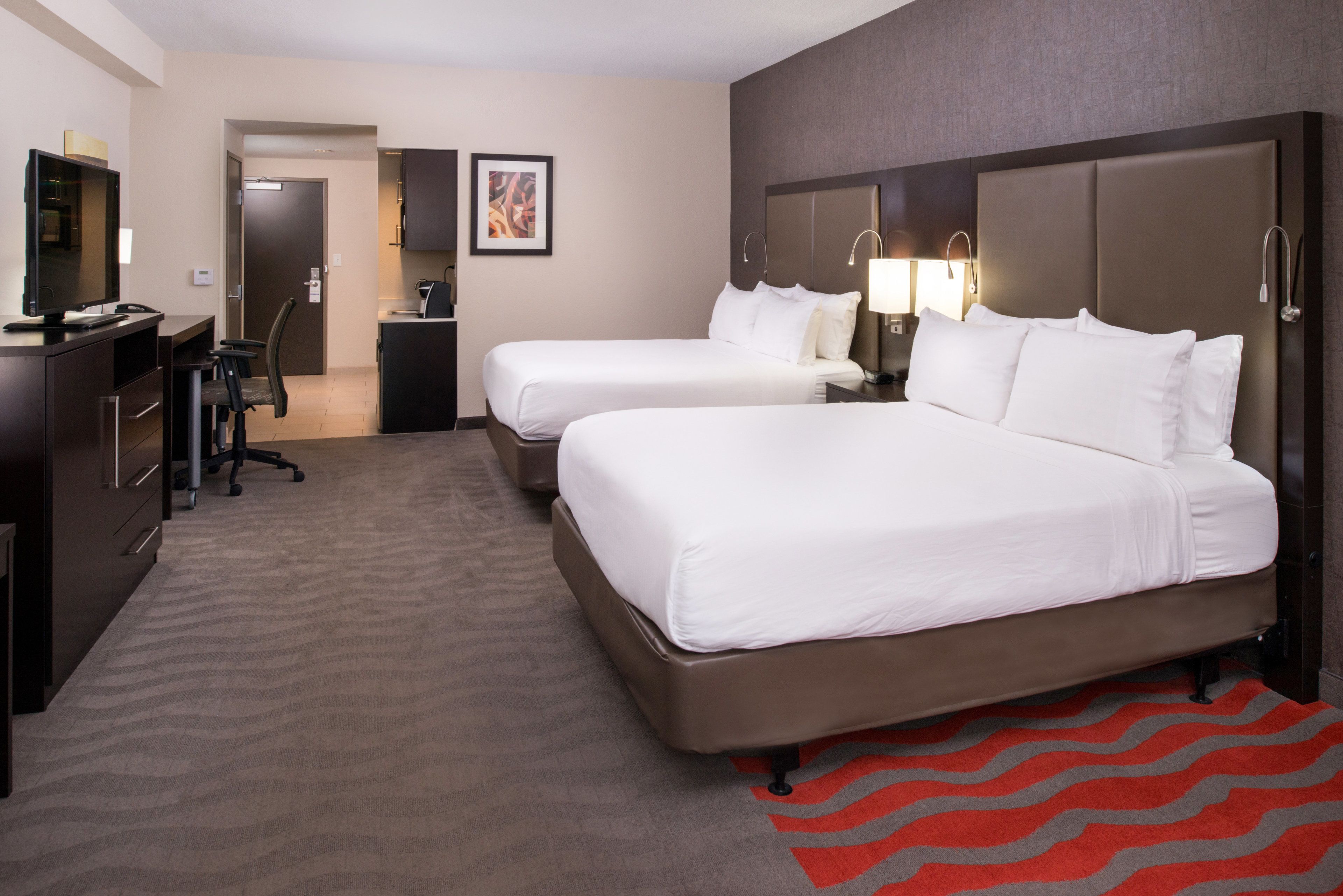 A hotel room with two beds, a desk, a chair, a TV, and a red rug.