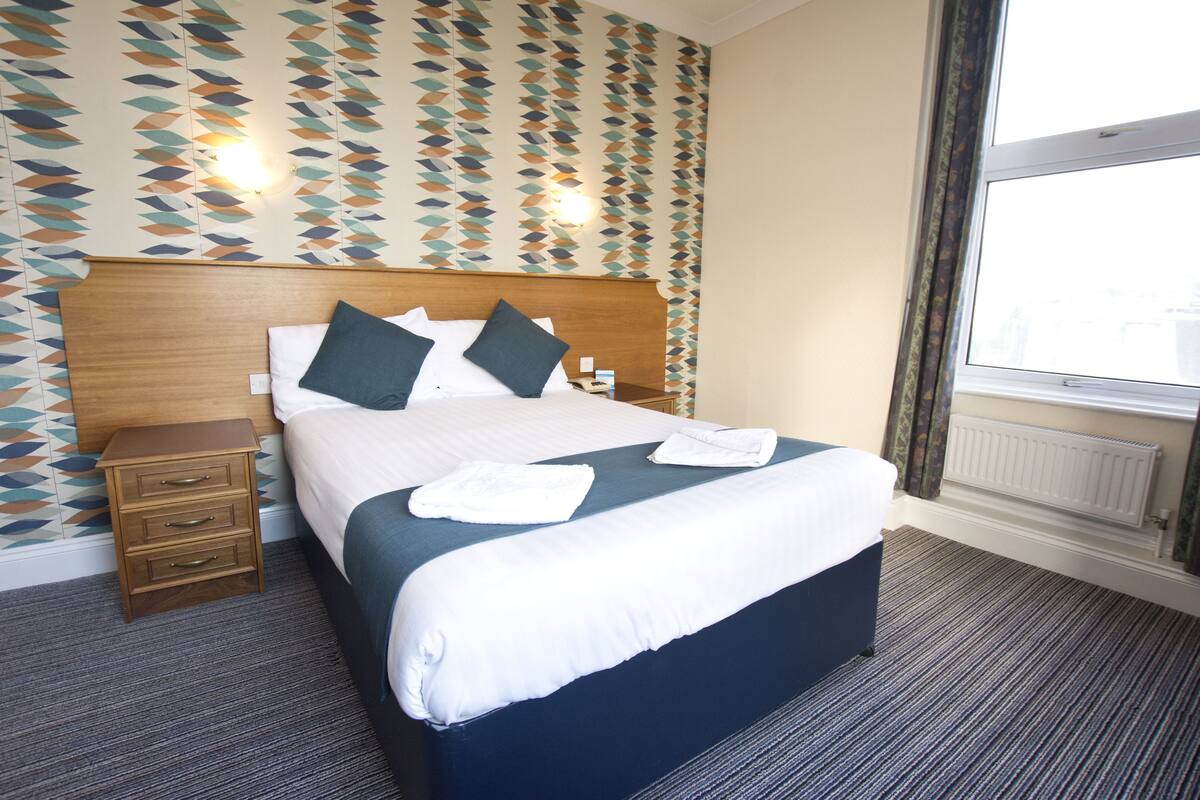 A neatly made bed with pillows and a folded towel, a wooden bedside table with drawers, and a patterned wall.