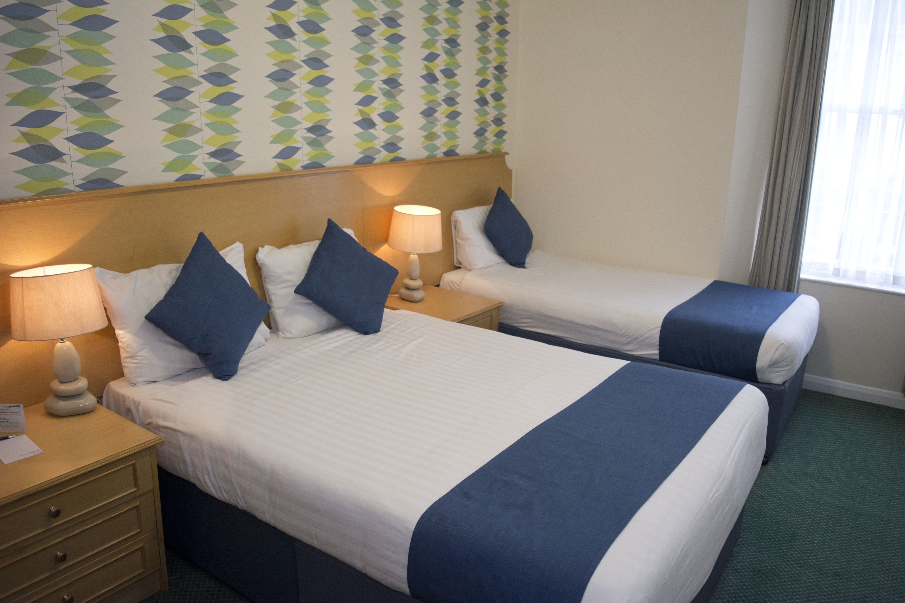 A hotel room with two beds, each with a blue accent pillow and a bedside lamp.