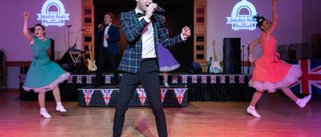 Un escenario con artistas en un ambiente formal, incluyendo un hombre con chaqueta de cuadros y dos mujeres con vestidos, con un fondo que muestra un logotipo y banderas británicas.
