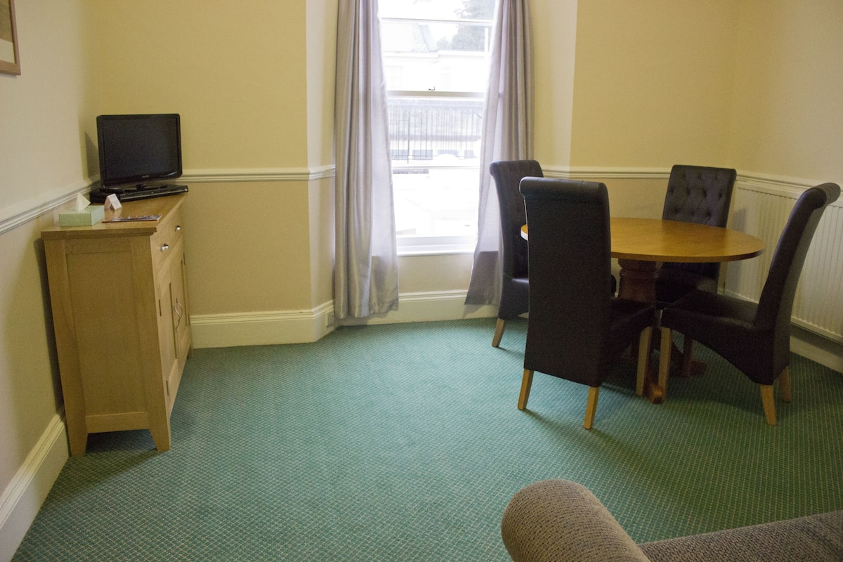 A room with a wooden cabinet, a television, a dining table with chairs, and a window with curtains.