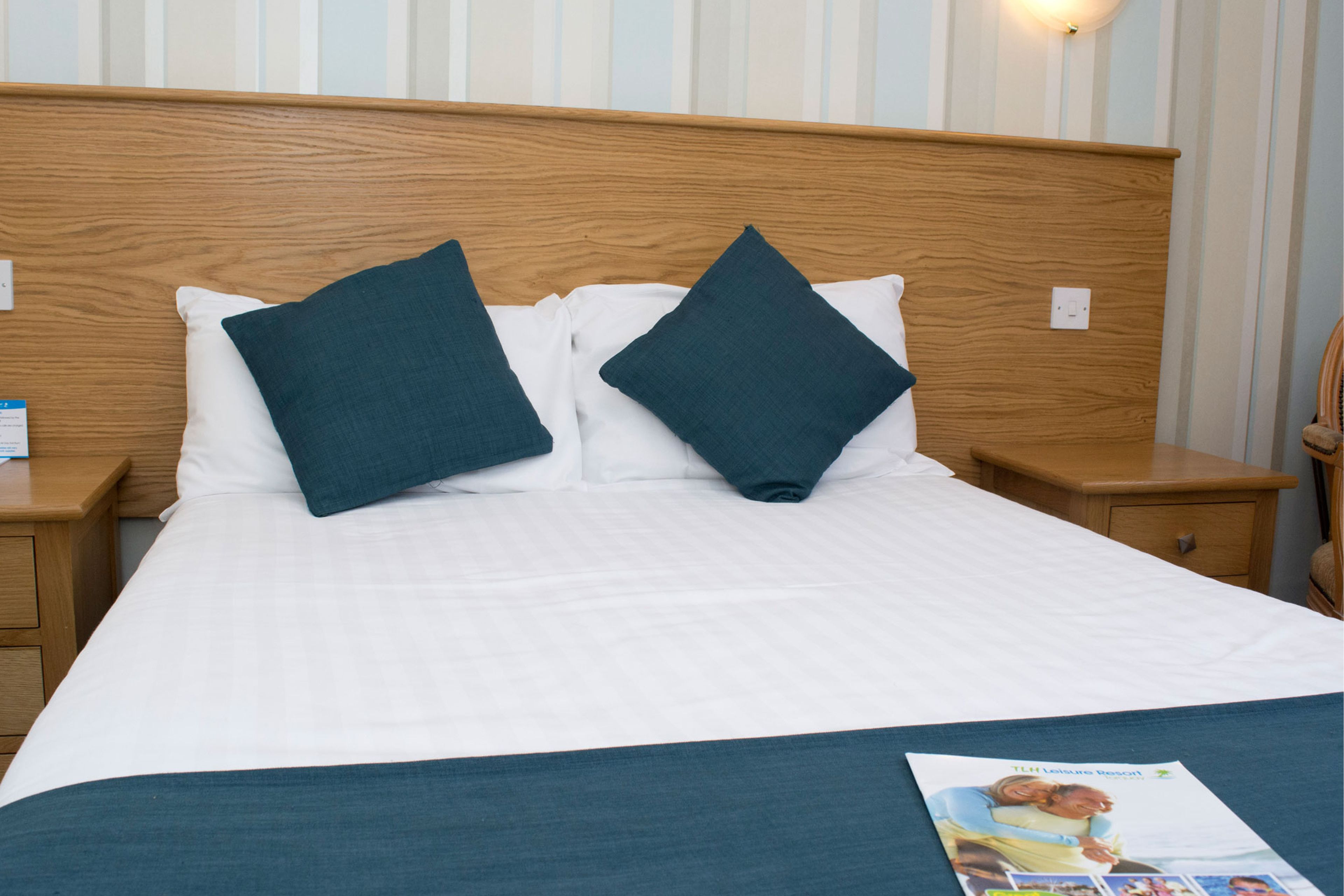 A neatly made bed with white bedding and two dark blue pillows, a wooden headboard, and a nightstand with a lamp on the left.