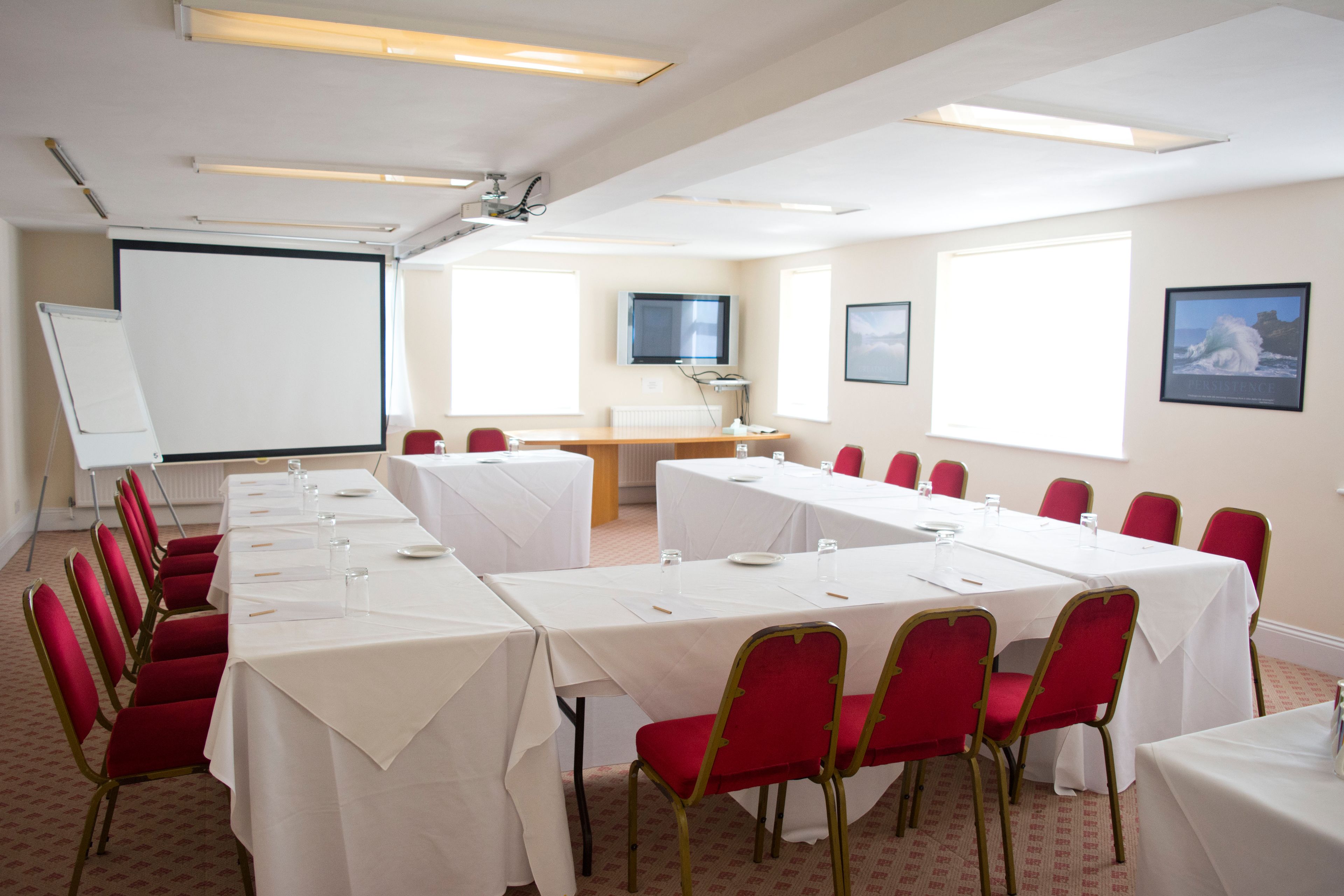 A conference room with long tables, red chairs, a whiteboard, and a projector screen.