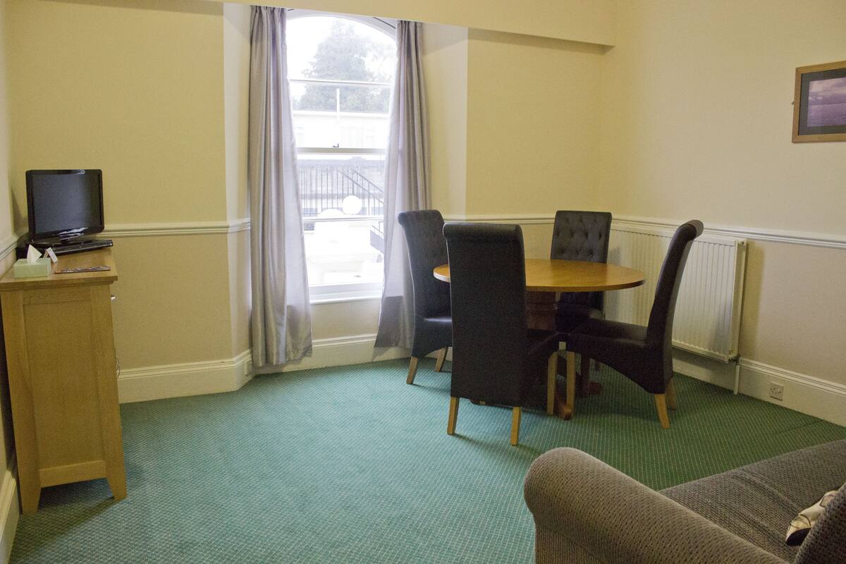 A room with a dining table, four chairs, a television on a stand, and a sofa.