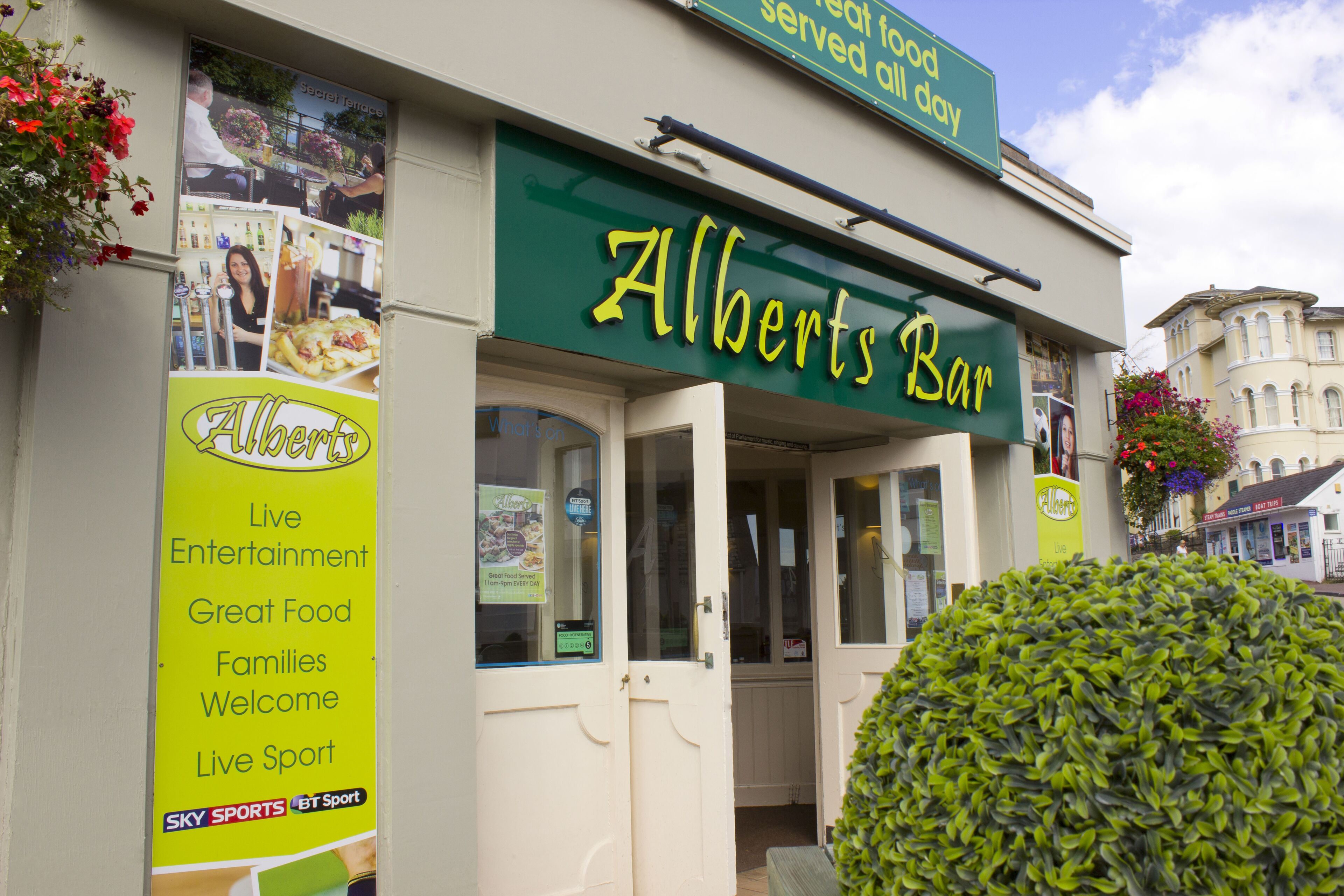 Un escaparate con un cartel que dice "Albert's Bar" y un banderín que anuncia "Espectáculos en vivo, comida excelente, familias bienvenidas, deportes en directo."