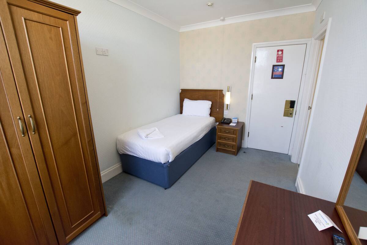 A hotel room with a bed, a wardrobe, a bedside table, and a door with a fire exit sign.