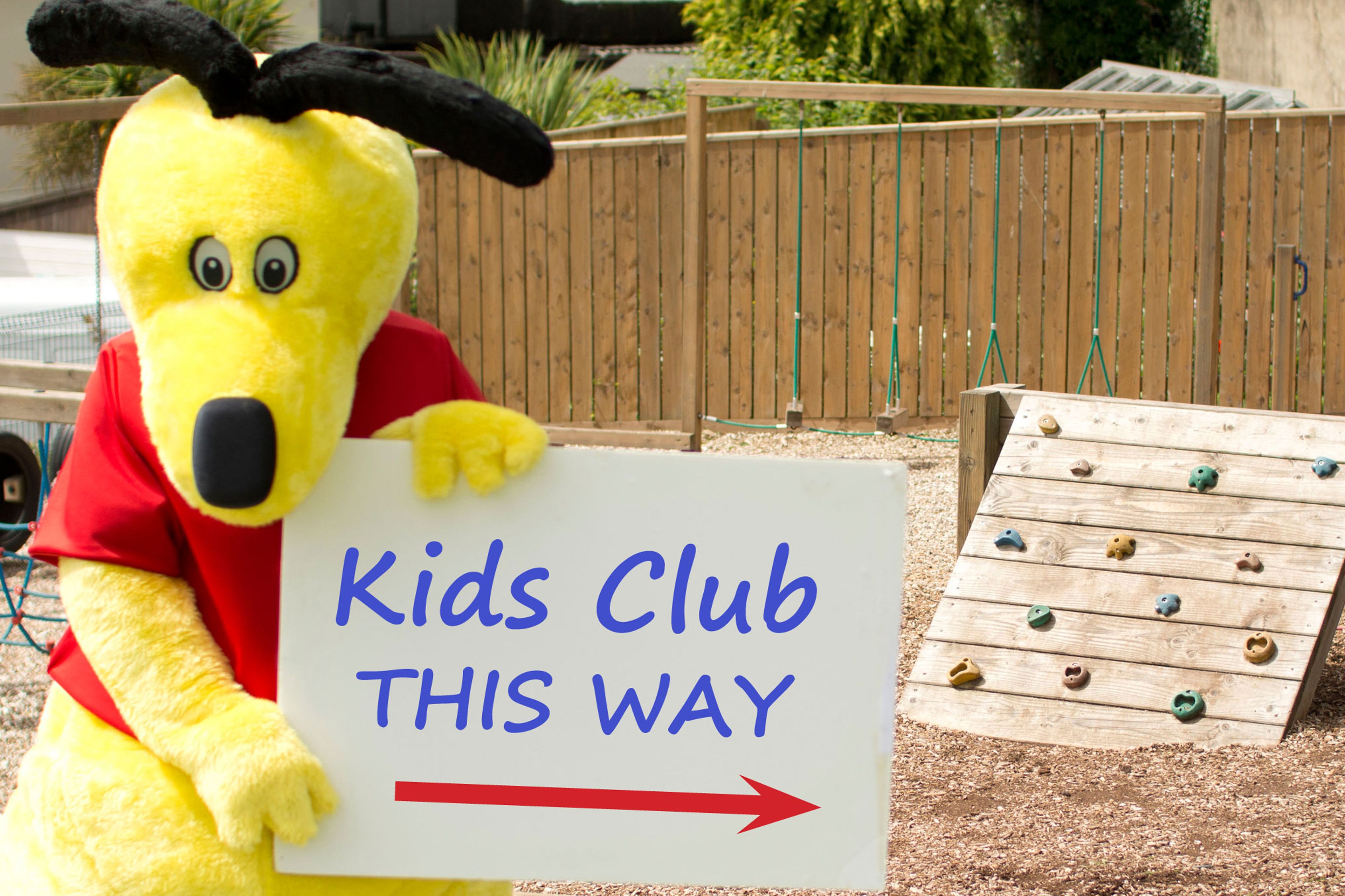 Un personaje promocional sostiene un letrero que dice 'Kids Club por aquí' con una estructura de escalada de madera cerca.