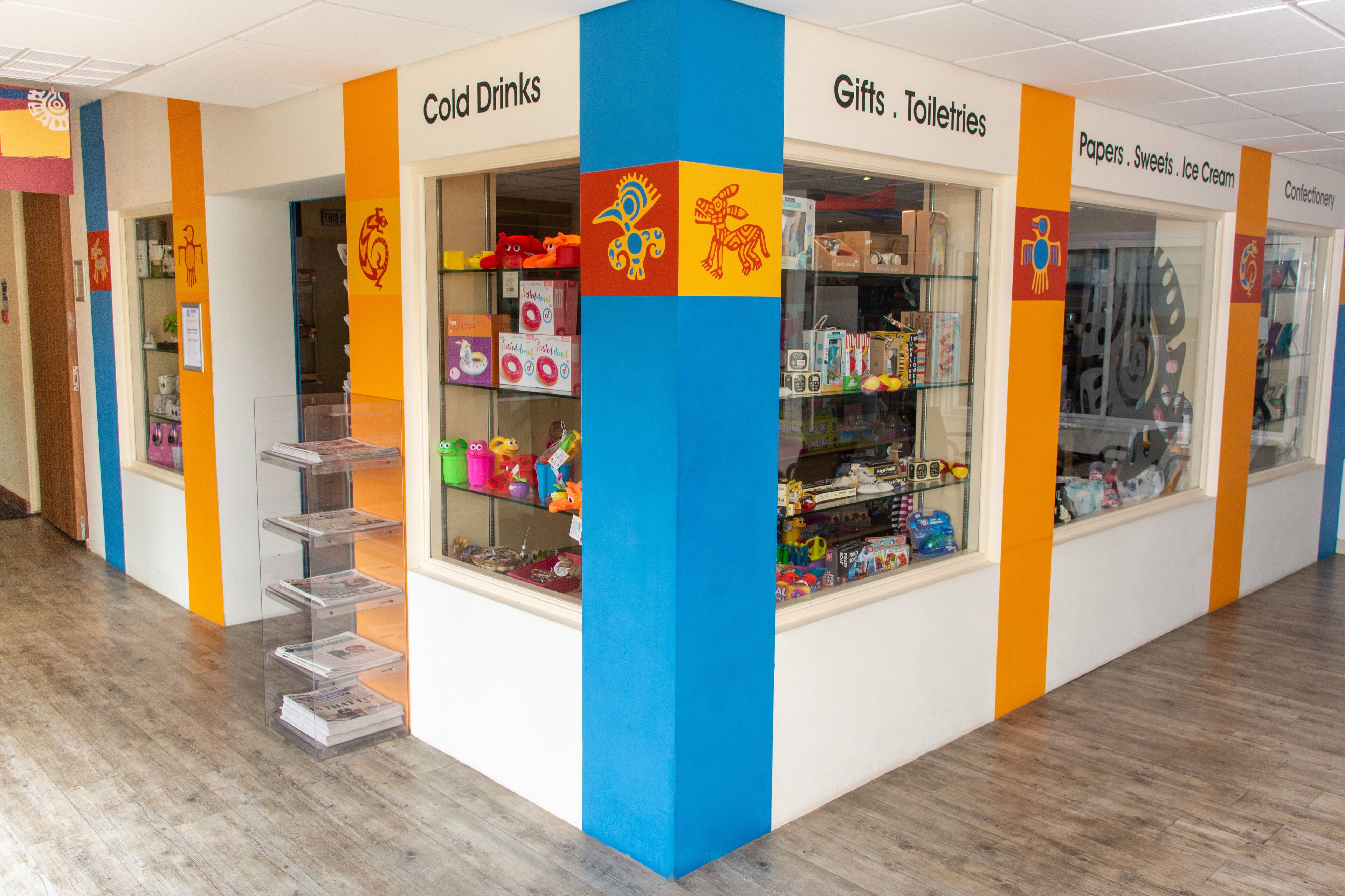 A brightly colored store with sections labeled 'Cold Drinks,' 'Gifts,' 'Toiletries,' and 'Confectionery.'