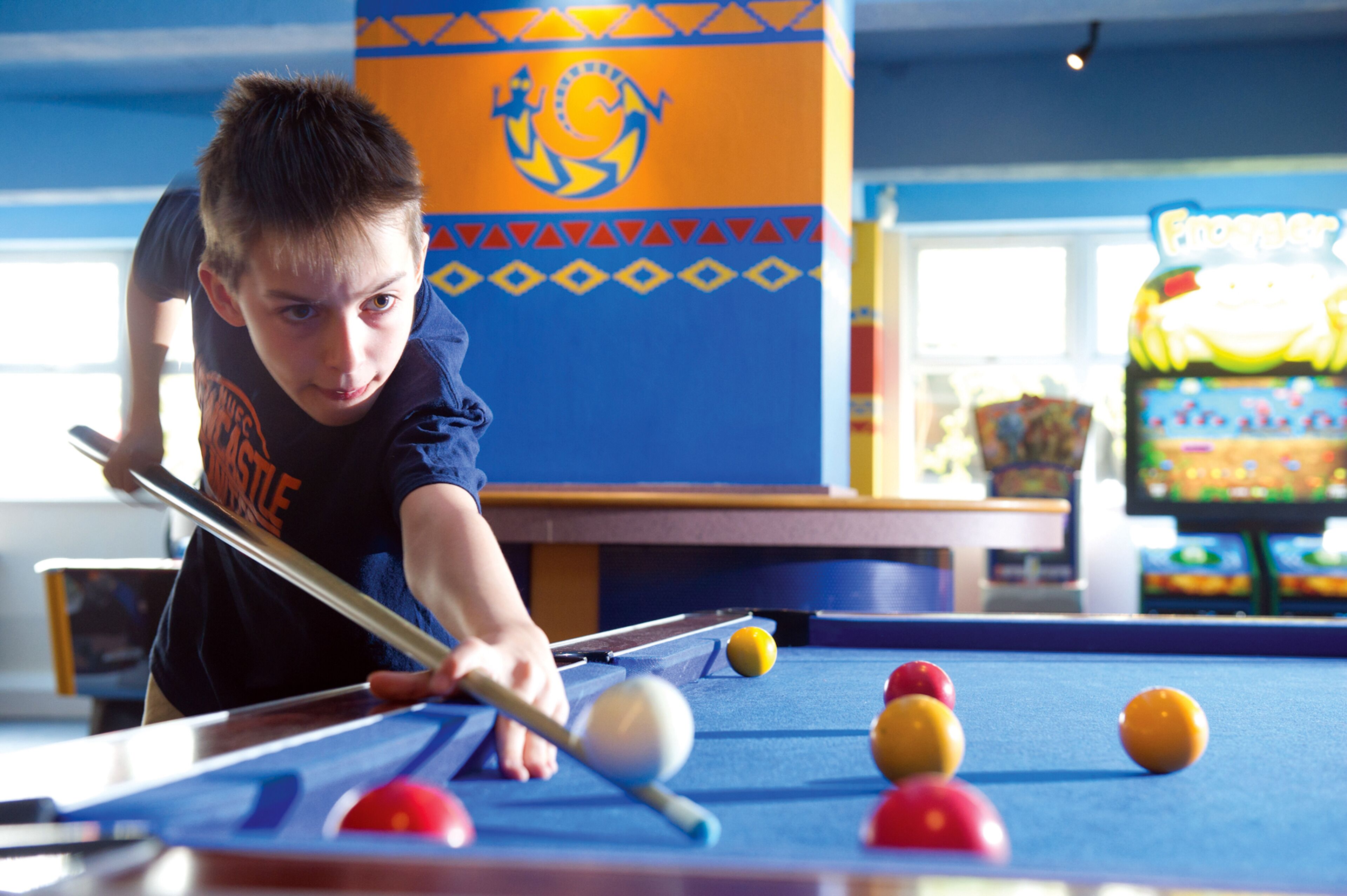 A pool table with balls and a cue stick, a colorful wall with a logo, and a visible arcade machine in the background.