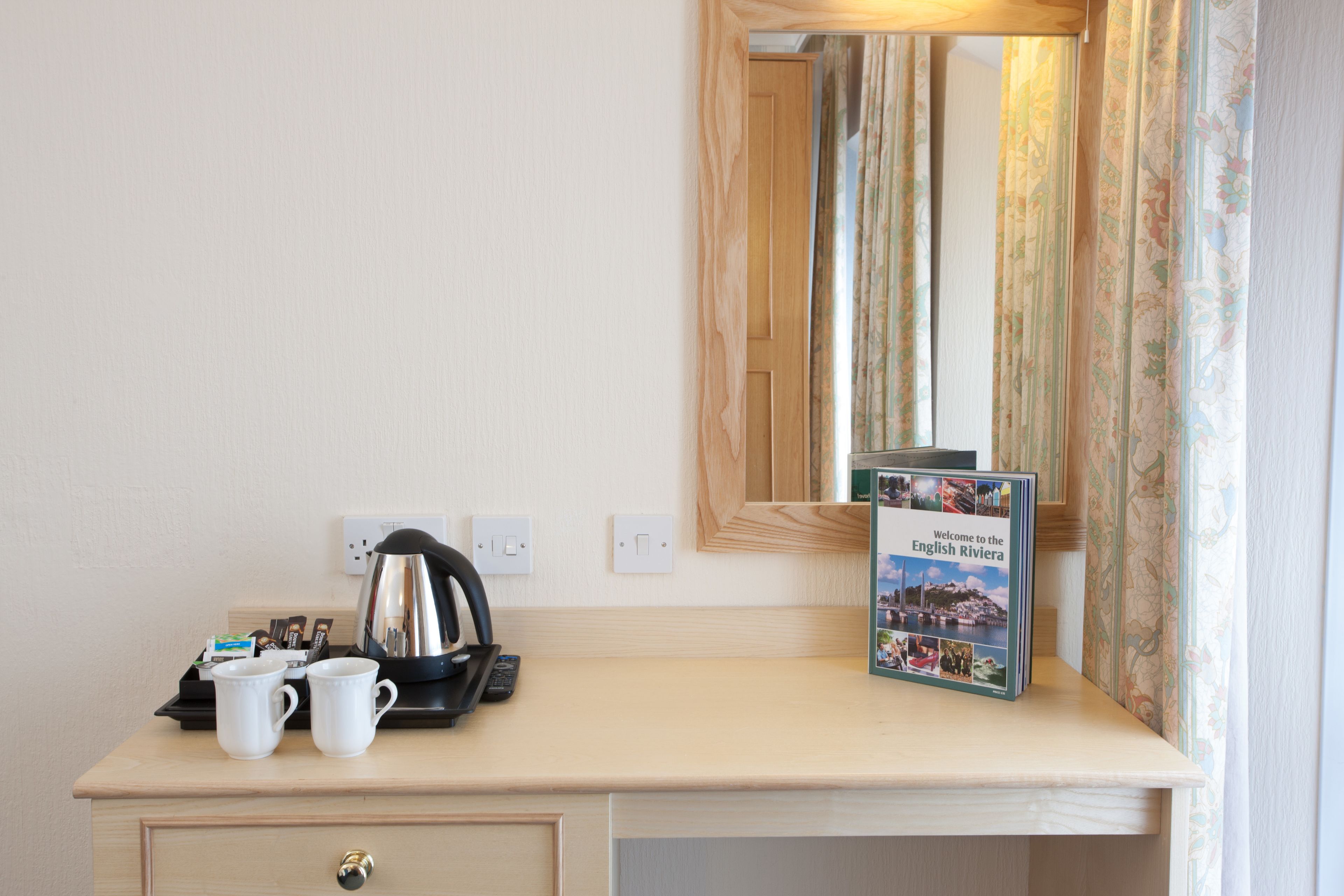 Een hotelkamer met een houten bureau, twee witte mokken, een waterkoker, een afstandsbediening en een welkomstfolder.