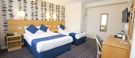 A hotel room with two beds, a desk, and a chair. There is a wall-mounted TV and a small shelf with a phone and a card.