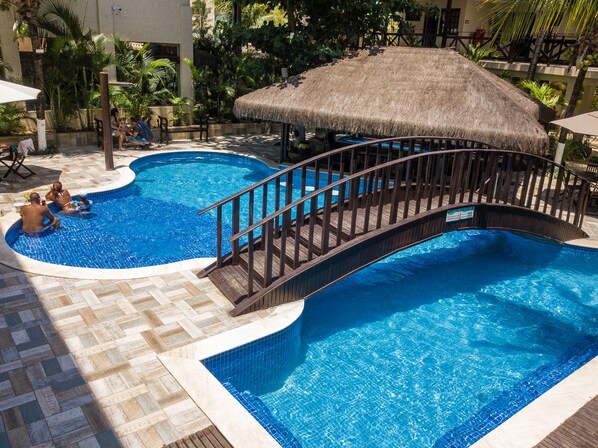 Una piscina con un puente de madera, rodeada de palmeras y una estructura con techo de paja.