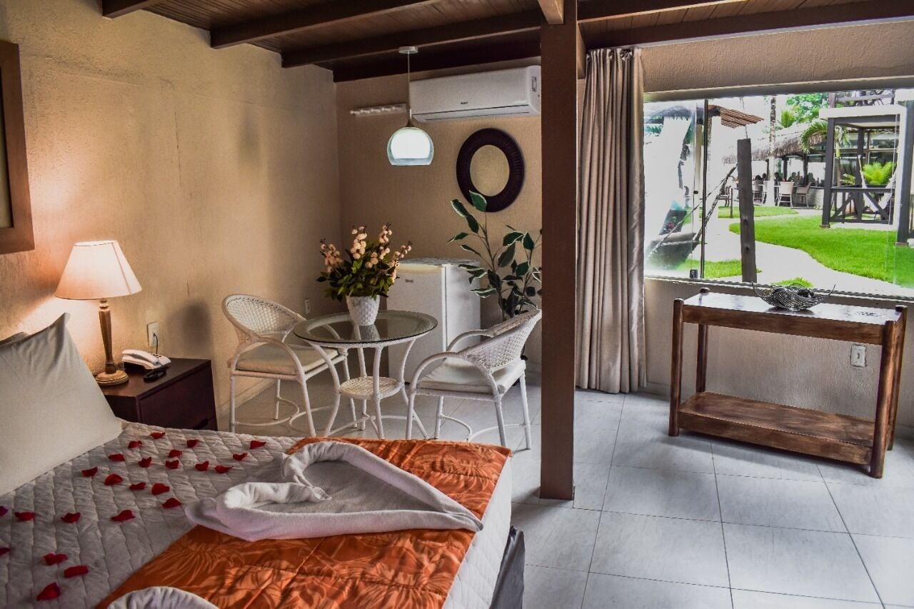 A hotel room with a bed, a small table with chairs, a window with a view of the garden, and a wall-mounted air conditioner.