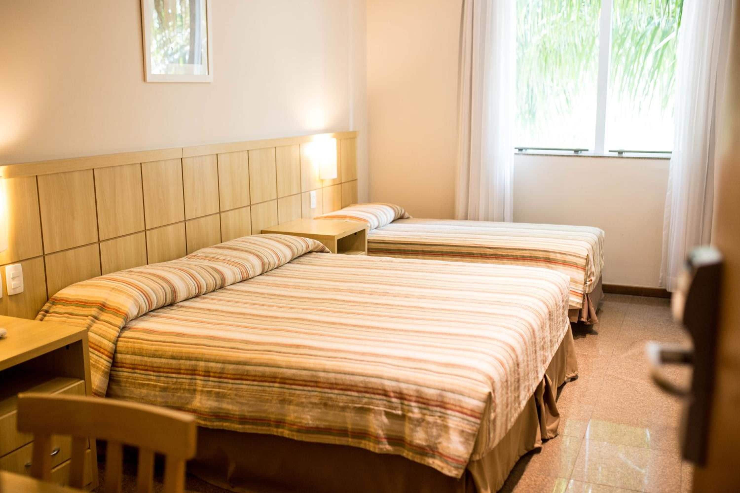 A hotel room with two beds, a wooden headboard, a bedside table, and a window with curtains.