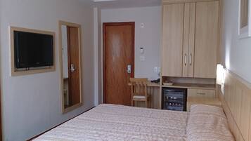 Un dormitorio con cama, televisor montado en la pared, armario de madera y un pequeño escritorio con silla.
