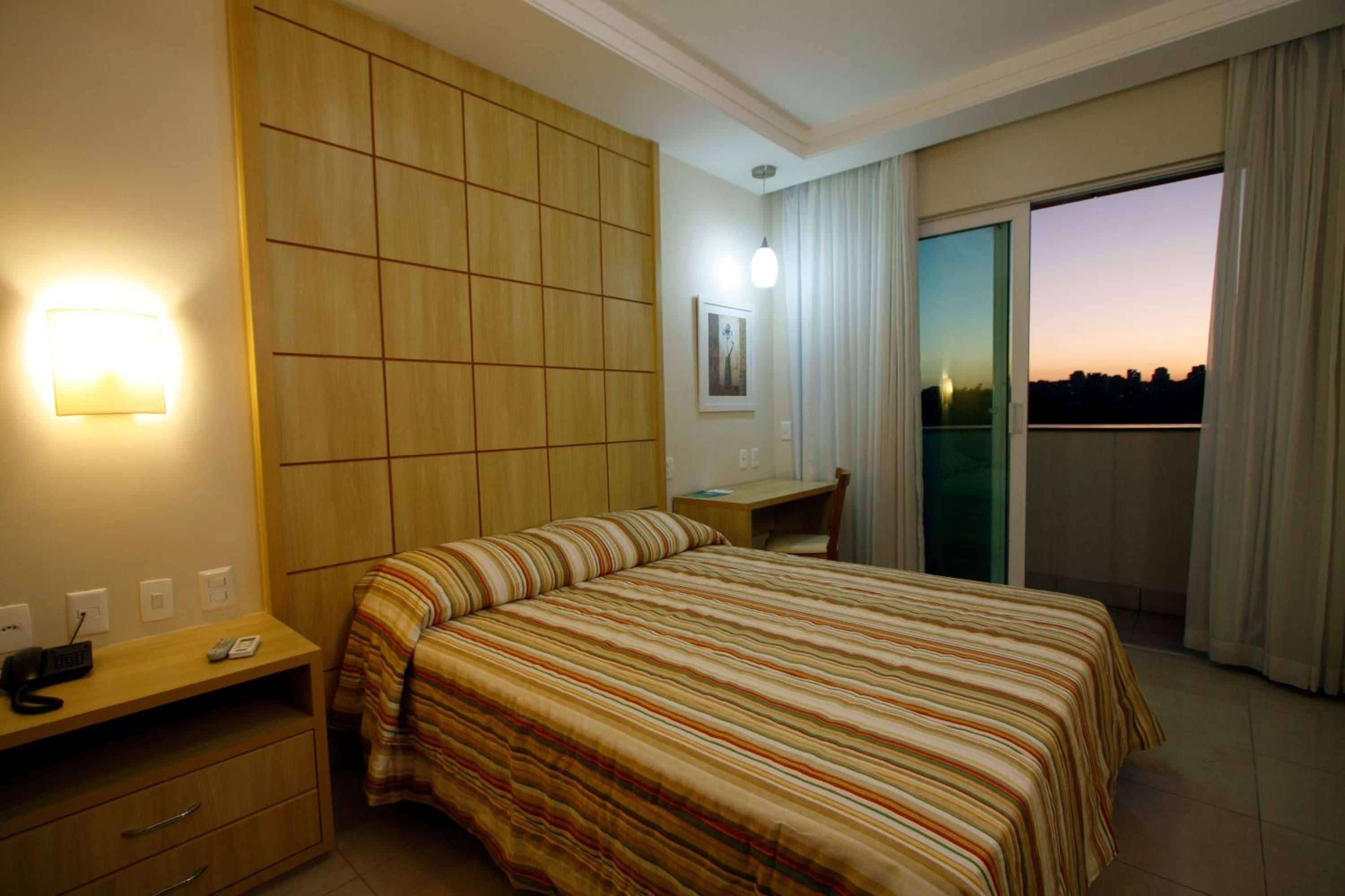 Une chambre d’hôtel avec un lit, un bureau, une chaise, un téléphone et une vue sur la ville au coucher du soleil.
