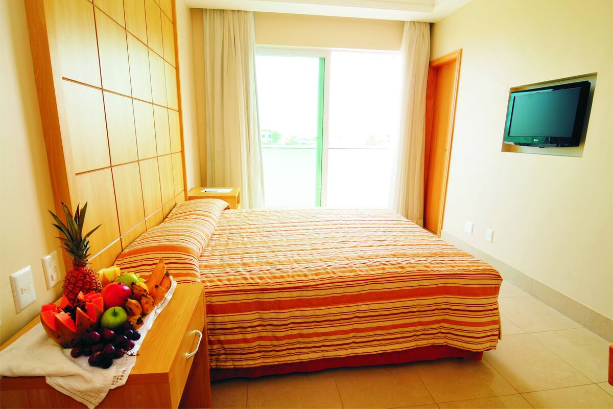 Une chambre d’hôtel avec un lit, un téléviseur fixé au mur et un panier de fruits sur la table de chevet.