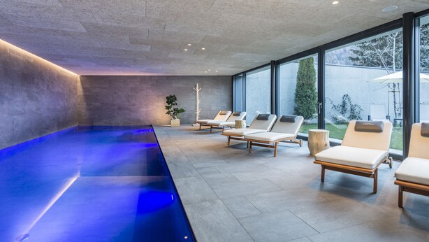A modern indoor pool area with a blue-lit water feature, lounge chairs, and large windows offering a view of the outdoors. - Lener Wirtshaus & Hotel (Campo di Trens)