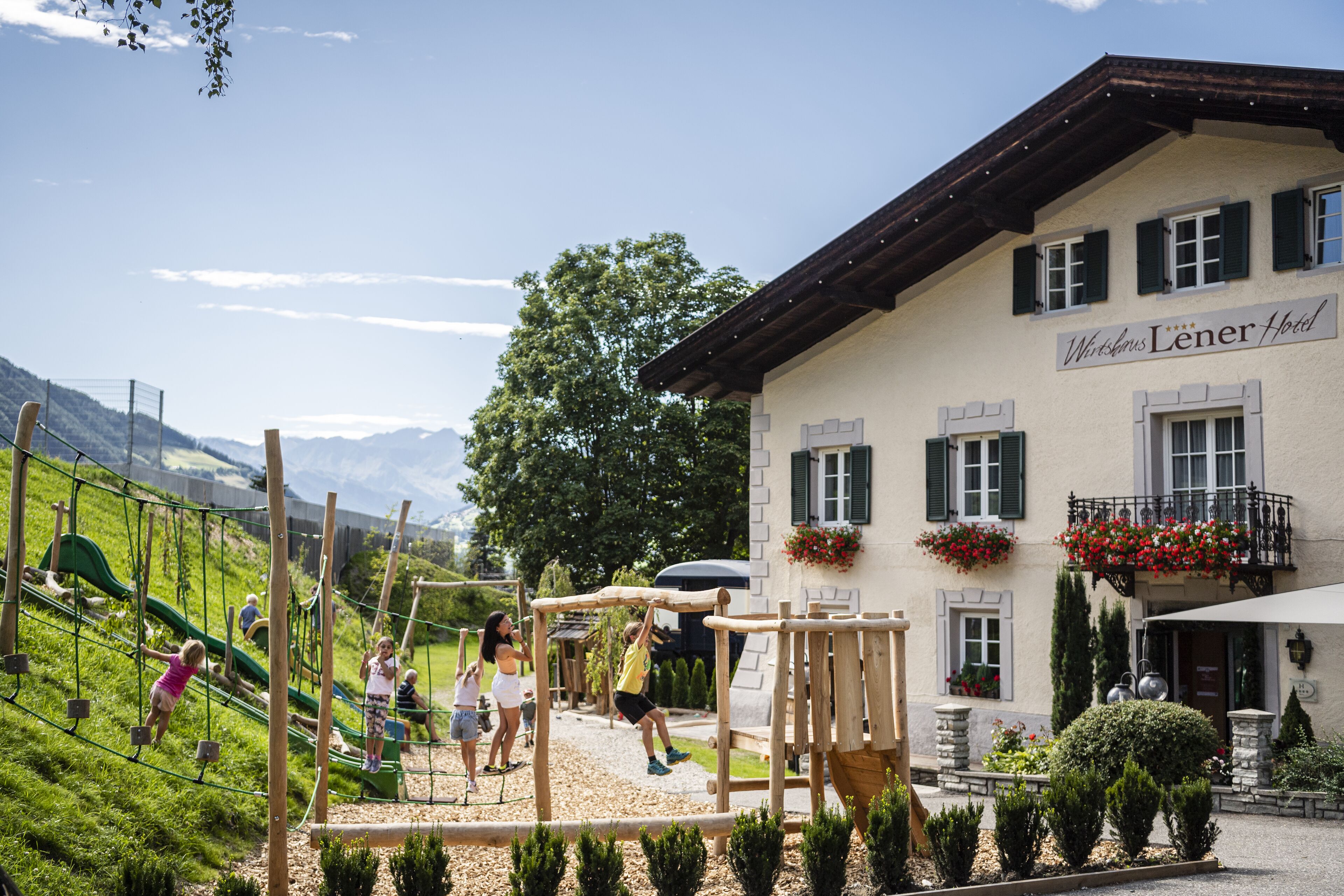 Un'area giochi con bambini che giocano, un edificio con il cartello che recita “Wiener Lener Hotel” e un paesaggio montuoso sullo sfondo.