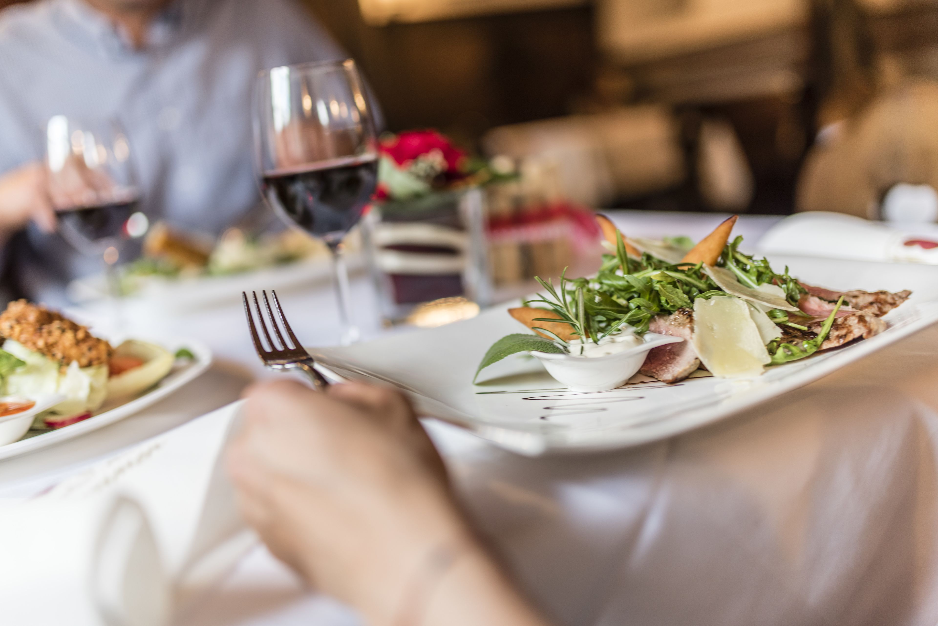 A dining table with plates of food, wine glasses, and a person holding a fork.