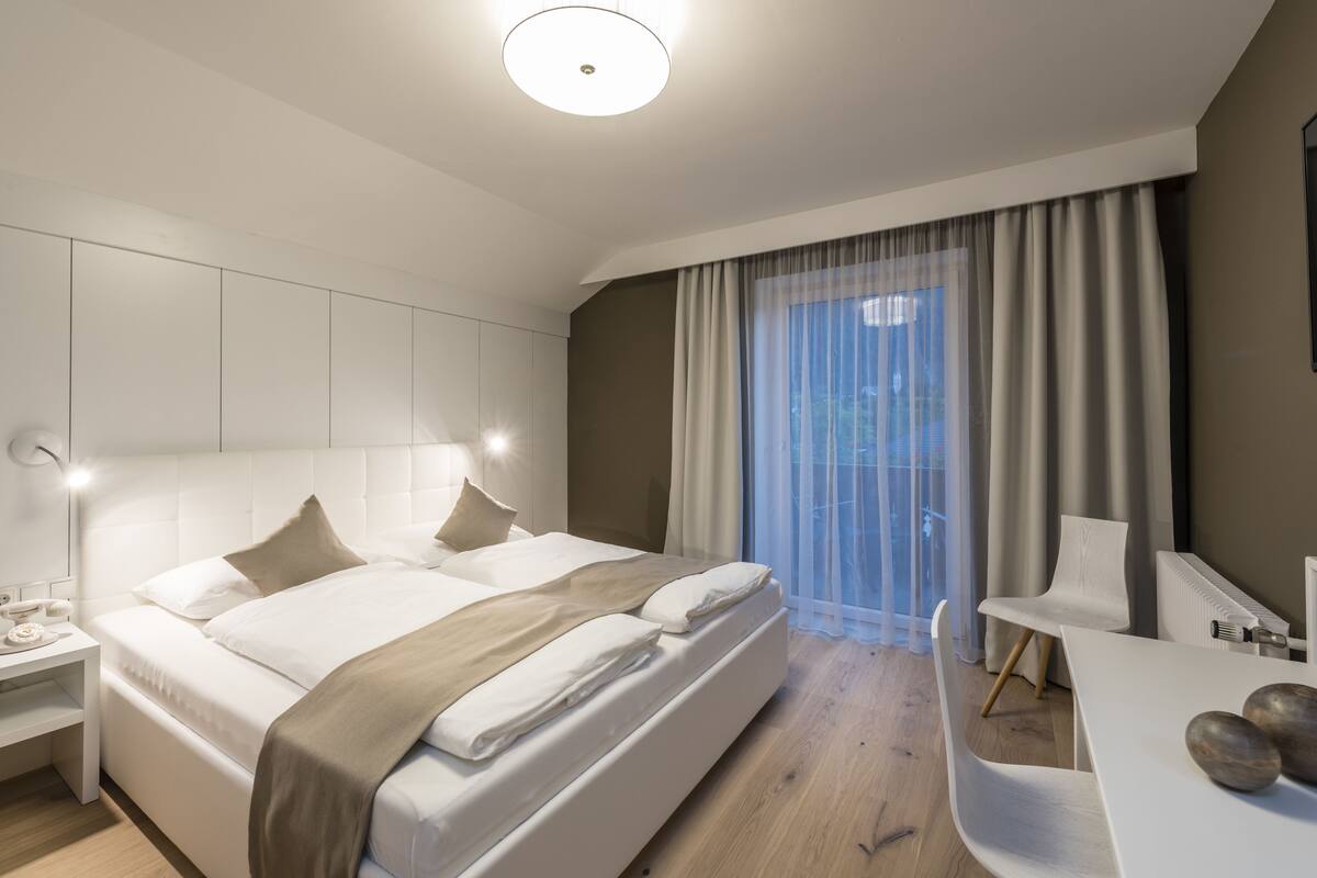 A modern hotel room with a large bed, bedside tables, a desk, and a view of the outdoors through sheer curtains.