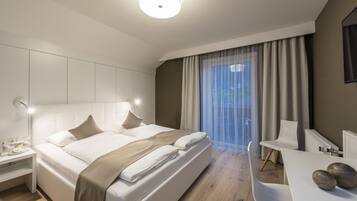 Quarto de hotel moderno com uma cama grande, mesas de cabeceira, uma escrivaninha e vista para o exterior através de cortinas finas.