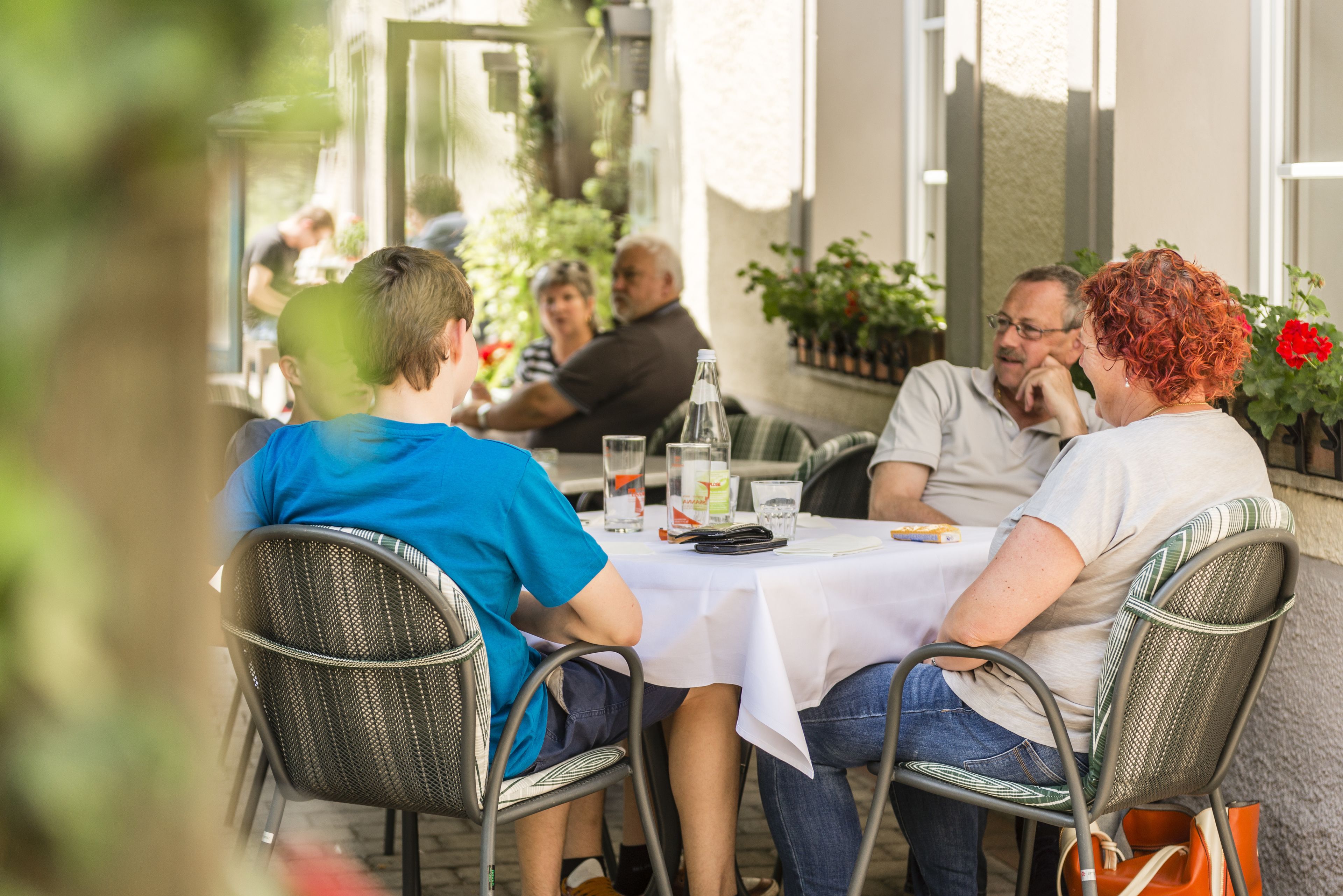 Un grupo de personas cenando en una mesa al aire libre con mantel blanco, rodeado de plantas en macetas y una fachada de edificio.